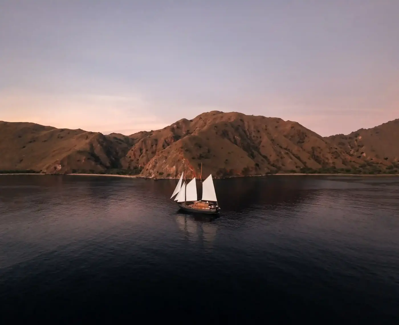 Phinisi at anchor beneath bare hills at dusk