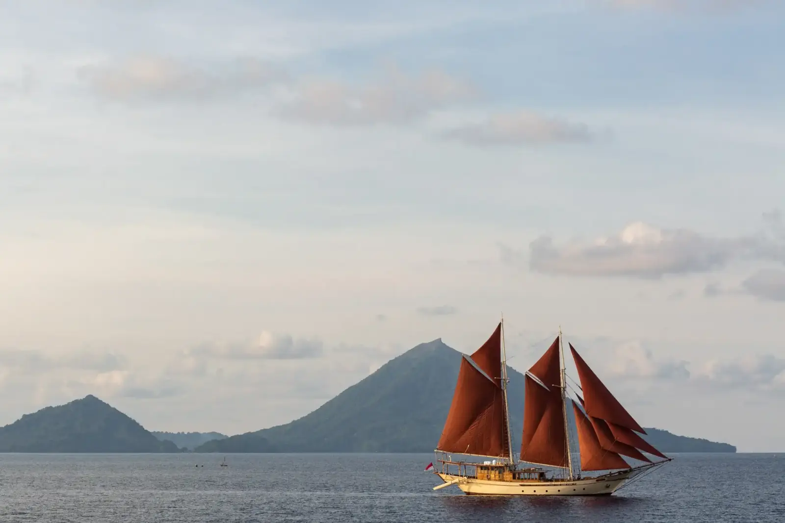 Phinisi with red sails below a volcano