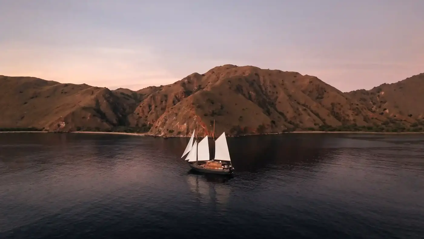 Phinisi at anchor beneath bare hills at dusk