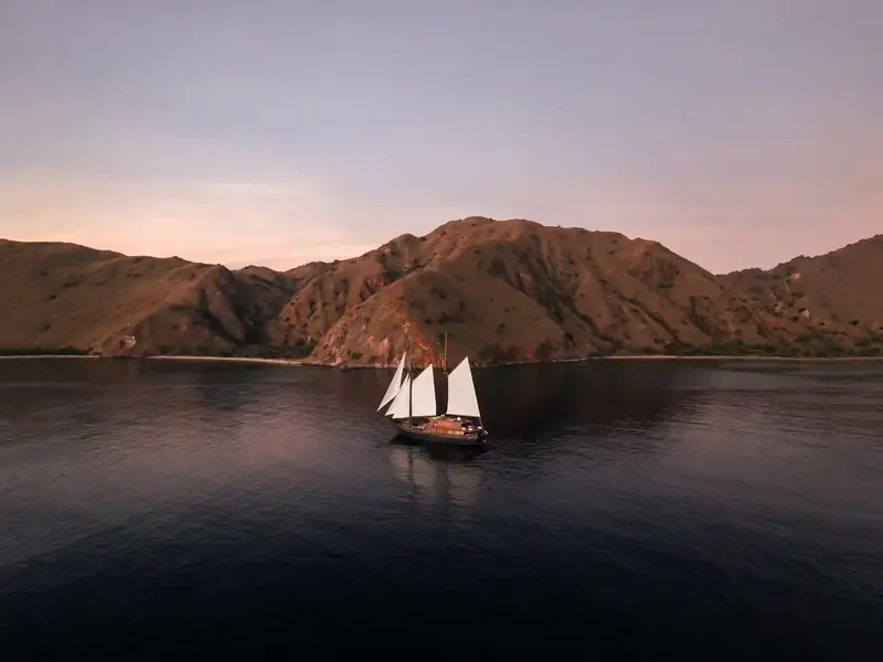 Phinisi at anchor beneath bare hills at dusk