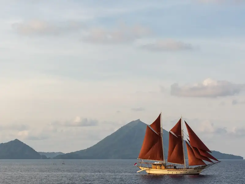 Phinisi with red sails below a volcano