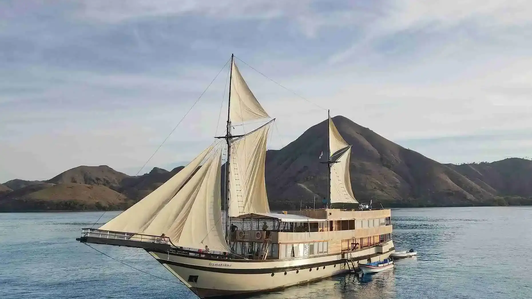 Phinisi with cream sails at anchor before green hills